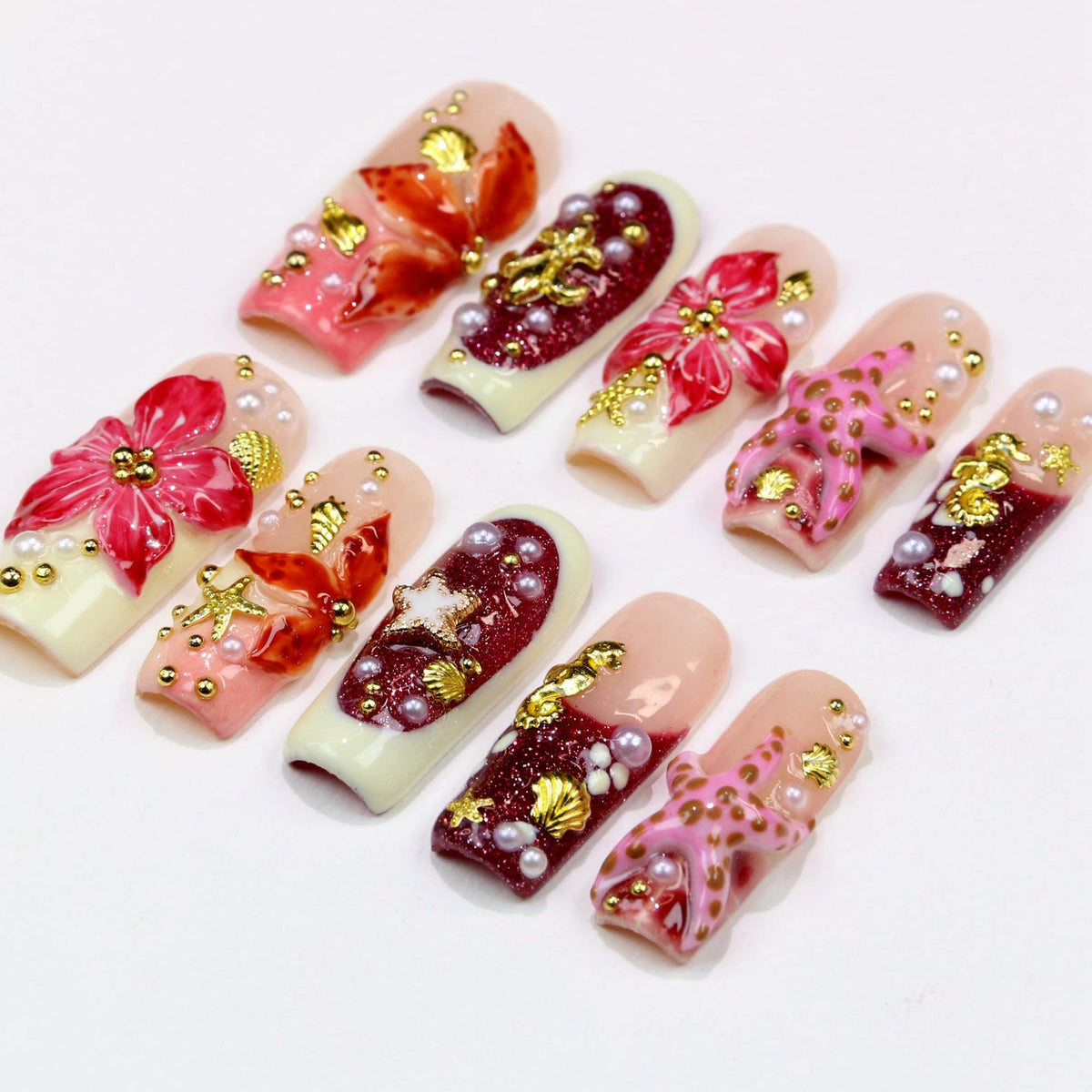 A collection of ten artificial nails, each with a unique design. The nails feature a variety of colors, including pink, red, and gold, with embellishments such as flowers, pearls, and starfish. The nails are arranged on a white surface.
