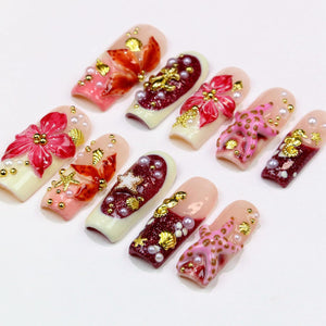 A collection of ten artificial nails, each with a unique design. The nails feature a variety of colors, including pink, red, and gold, with embellishments such as flowers, pearls, and starfish. The nails are arranged on a white surface.