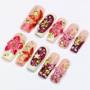 A collection of artificial nails with various designs. Some nails feature 3D floral and seashell embellishments in shades of pink, red, and gold. Others have glitter accents and pearl-like beads. The nails have a natural, nude base.