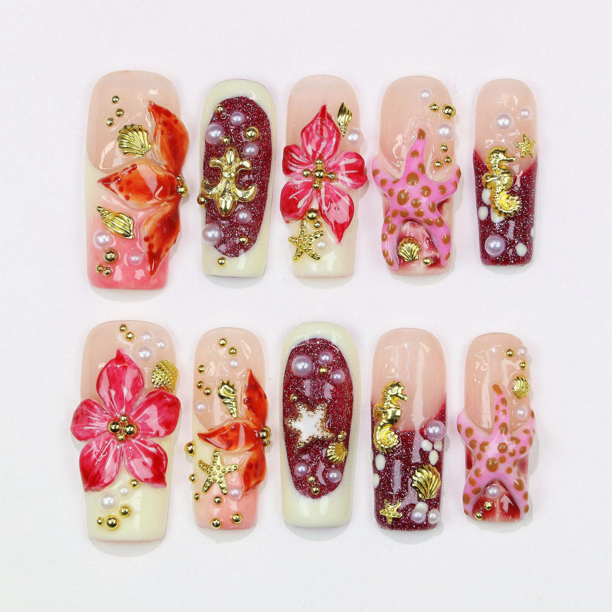 A set of ten artificial nails with a variety of designs. The nails feature a light pink base with red, gold, and white accents. Designs include flowers, starfish, seashells, and pearls.