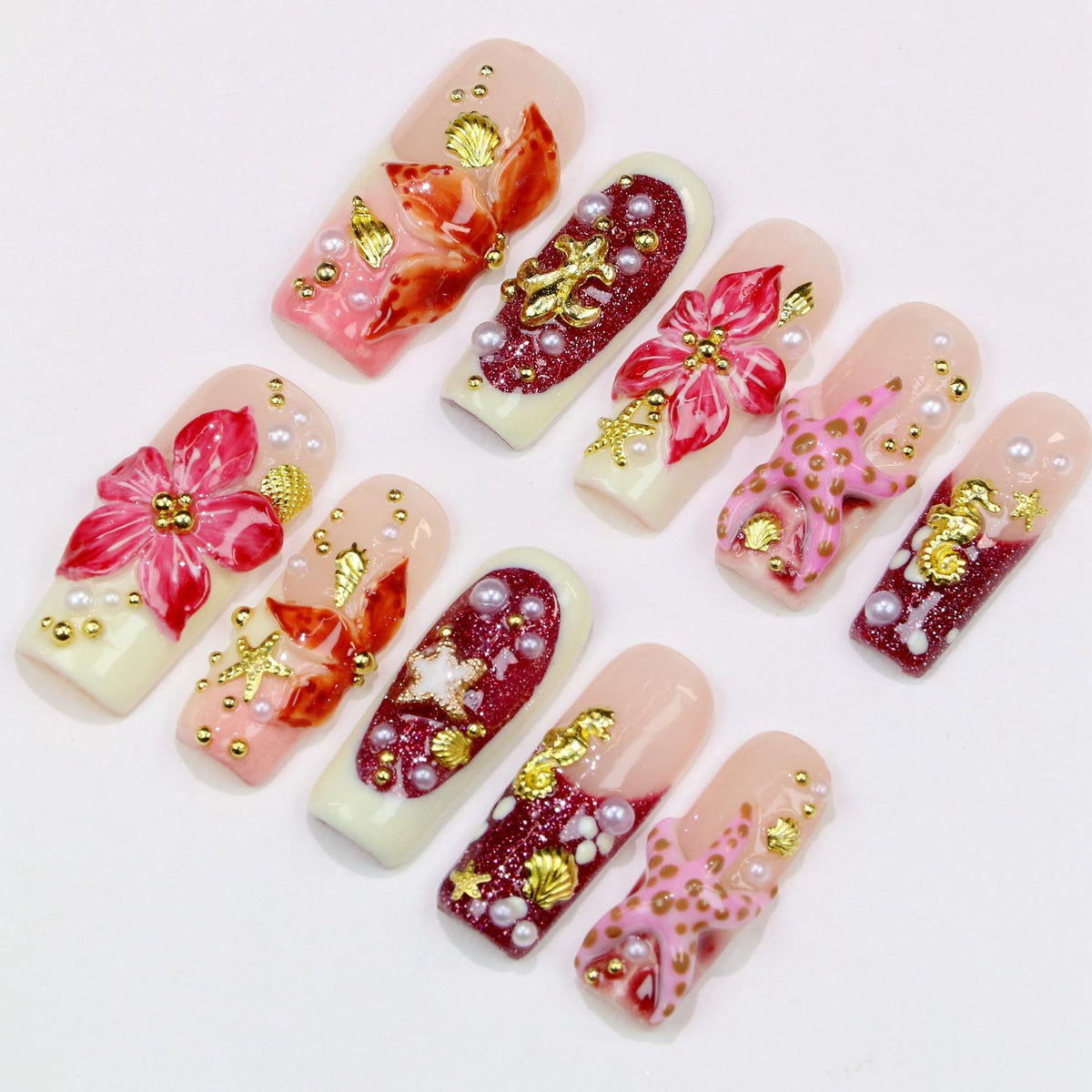 A collection of ten artificial nails, each with a unique design. The nails feature a variety of colors, including pink, red, and gold, with embellishments such as pearls, flowers, and seashell accents. The nails are arranged on a white surface.