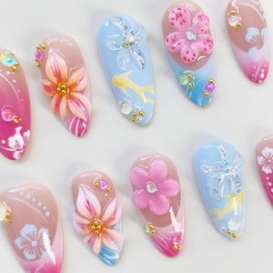 A collection of artificial nails with various designs. Some nails feature 3D flowers in pink and orange, while others have aquatic themes with yellow fish and glittery starfish on a blue background. Accents include small gold beads and iridescent gem