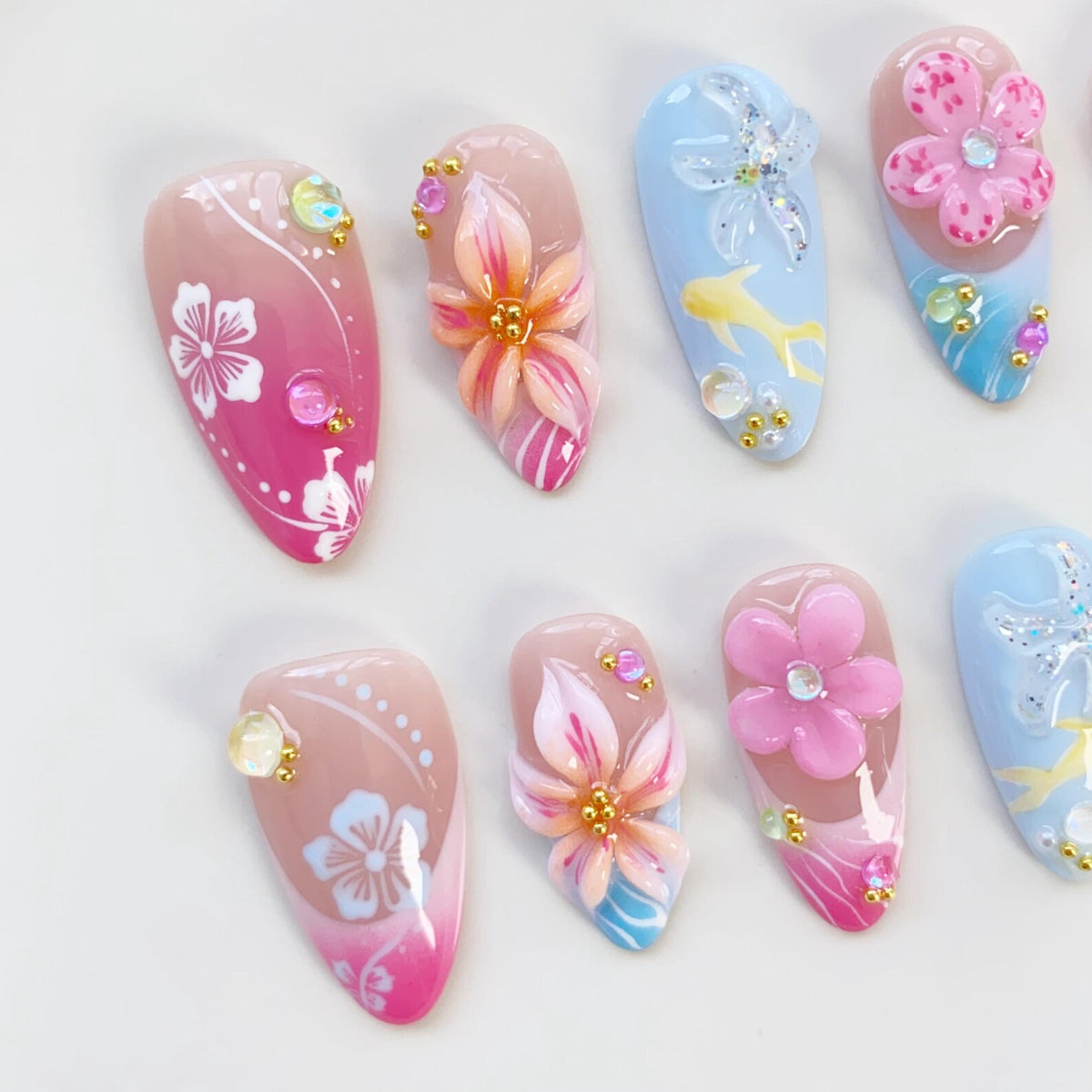 A collection of almond-shaped artificial nails with various designs. Some nails feature 3D flowers in pink and orange, others have painted flowers, and some have a blue background with a fish and starfish design. Embellishments include rhinestones an