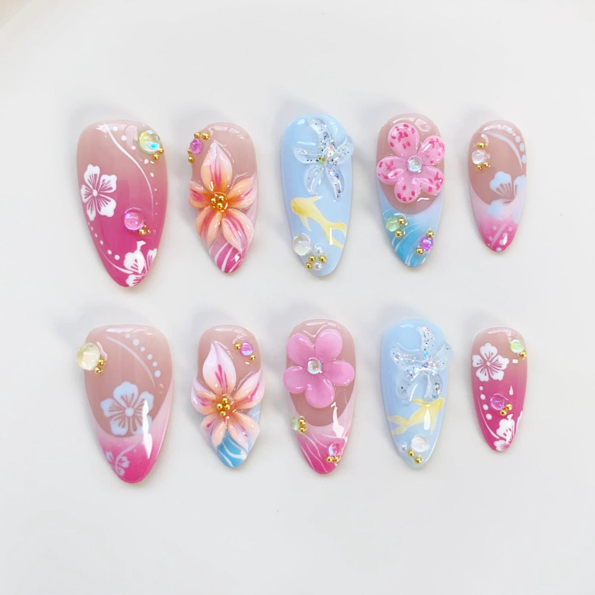 A set of ten artificial nails with floral designs. The nails feature a variety of colors, including pink, blue, and white, with 3D flower embellishments and sparkling accents. Each nail has a unique design.