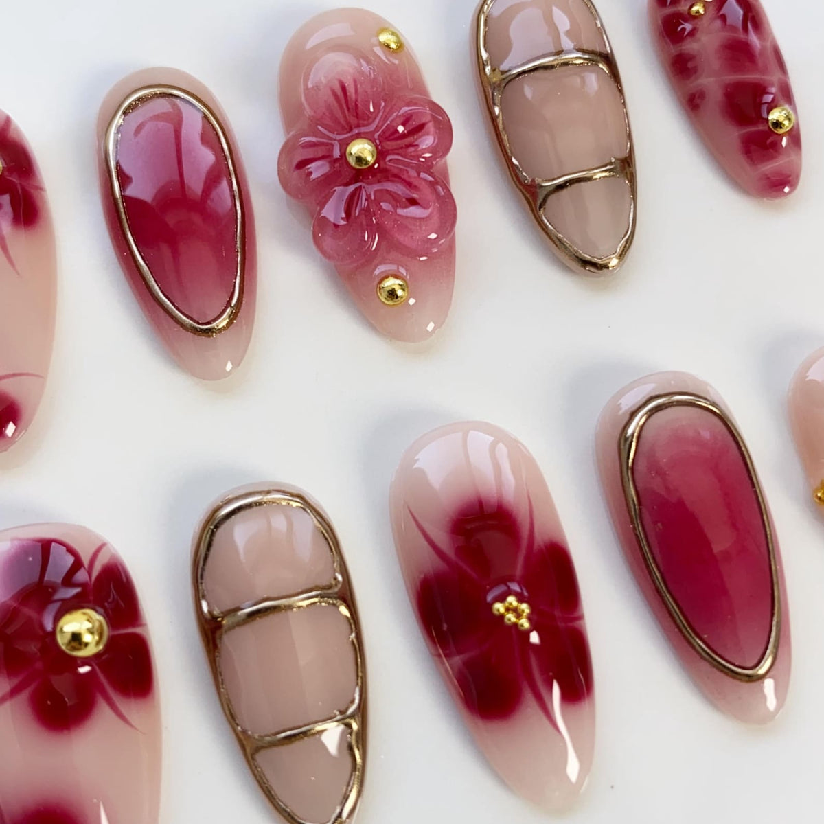A collection of artificial nails with various designs. Some nails feature a gradient of pink and red, while others showcase a 3D flower or floral patterns. Gold accents are present on several nails, adding a touch of elegance.