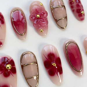 A collection of artificial nails with various designs. Some nails feature a gradient of pink and red, while others showcase a 3D flower or floral patterns. Gold accents are present on several nails, adding a touch of elegance.