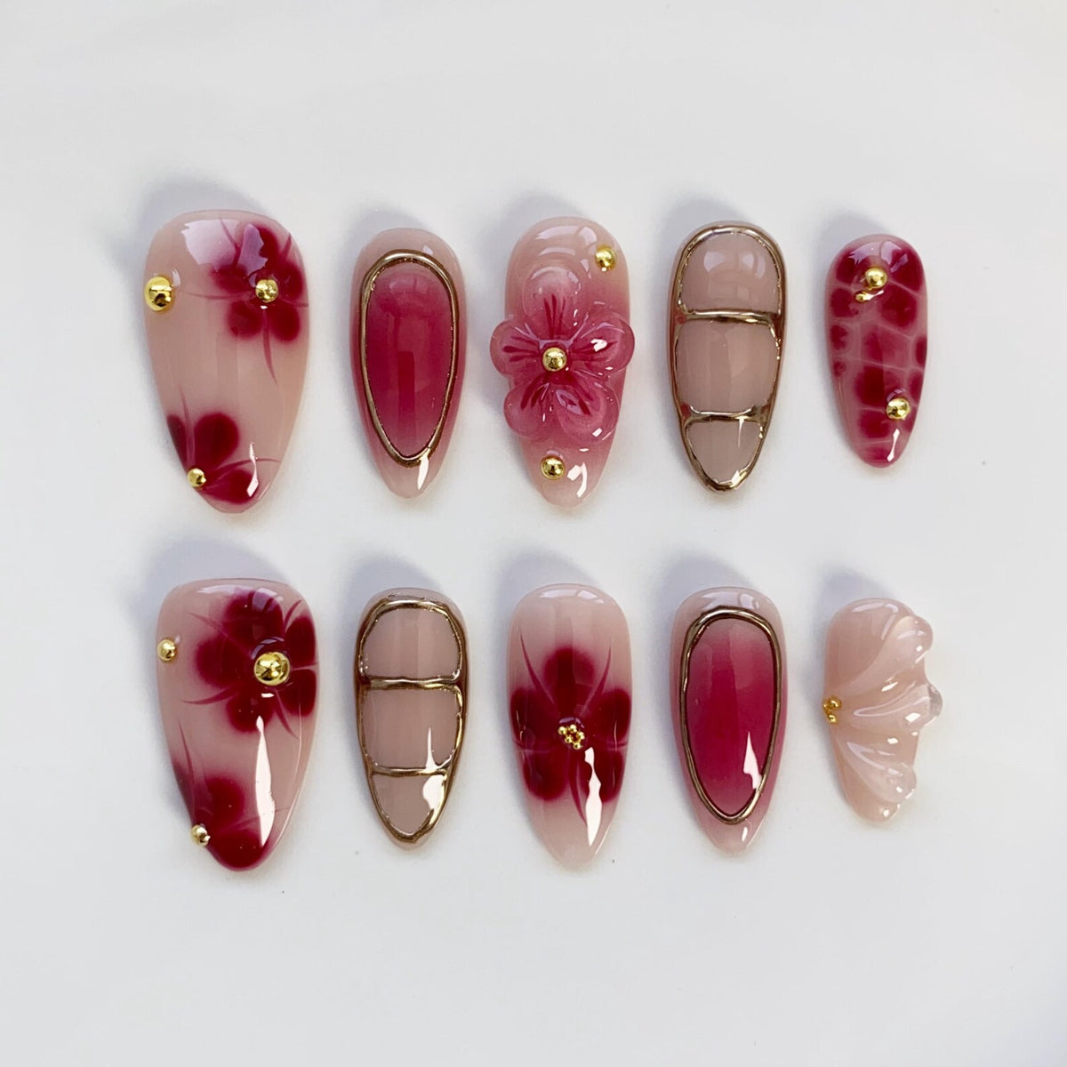 A set of ten artificial nails in various designs. The nails feature a combination of pink, red, and gold accents. Some nails have floral designs, while others have gold frames or small gold studs. The nails are oval-shaped.
