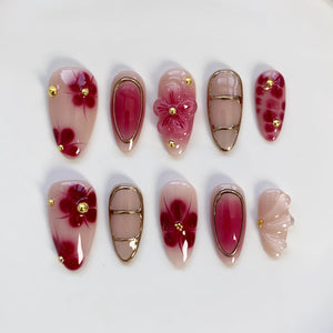 A set of ten artificial nails in various designs. The nails feature a light pink base with burgundy floral patterns, gold accents, and a 3D flower. Some nails have gold frames or solid burgundy designs.
