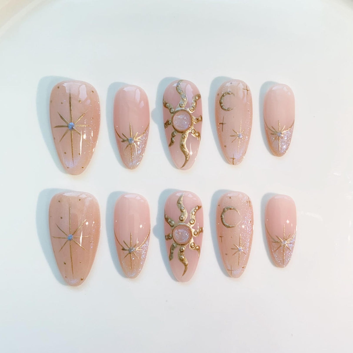 A set of ten almond-shaped press-on nails in a light pink hue. Each nail features gold celestial designs, including stars, suns, and moons, with glitter accents. The nails are arranged on a white surface.