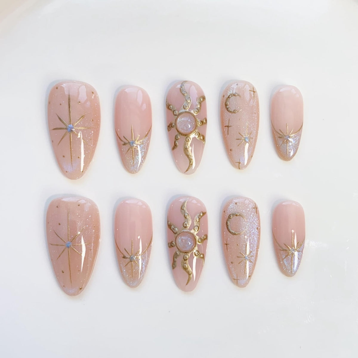 Set of ten almond-shaped false nails in a light pink hue. Each nail features celestial designs in gold and silver, including stars, suns, and moons. Some nails have a glittery accent at the base.