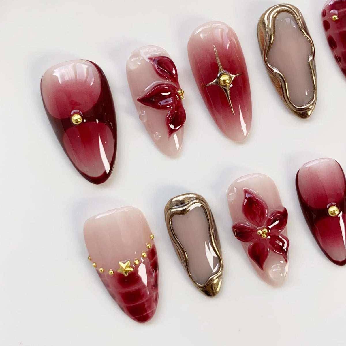 A collection of artificial nails with various designs. Some nails feature a gradient of red and pink hues, while others showcase 3D floral embellishments, gold star accents, and metallic gold frames. The nails are oval-shaped.