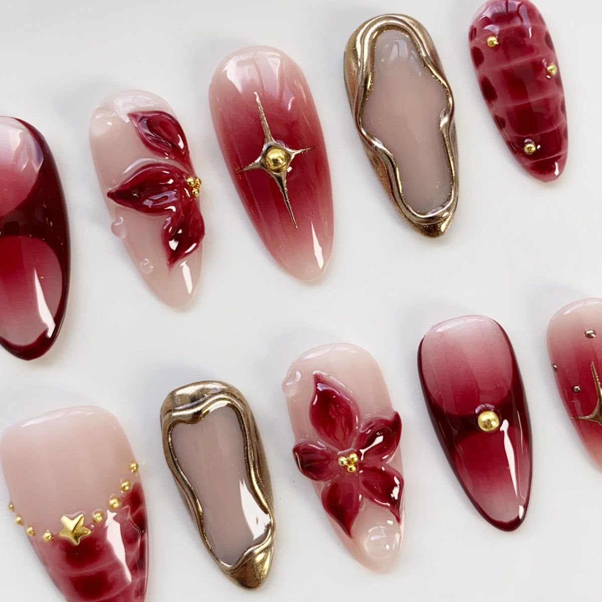 A collection of artificial nails with various designs. Some nails feature a gradient of red and pink hues, while others have gold embellishments, floral accents, and starburst patterns. The nails are oval-shaped and arranged on a white surface.