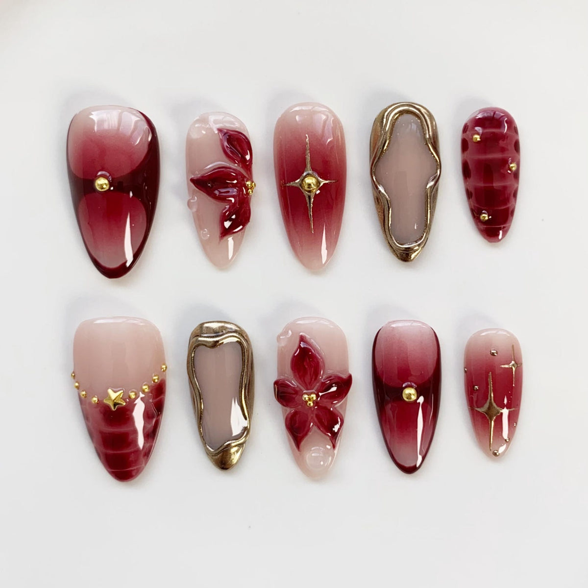 A set of ten artificial nails in various designs. The nails feature a mix of colors, including burgundy, gold, and nude. Some nails have floral or geometric embellishments, while others have a gradient effect.