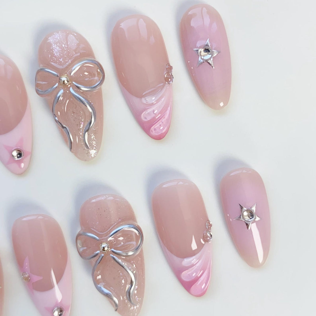 A set of almond-shaped artificial nails in various designs. Some nails feature a light pink base with silver bow accents, while others have a pink and white swirl design or a silver star. The nails are arranged on a white surface.