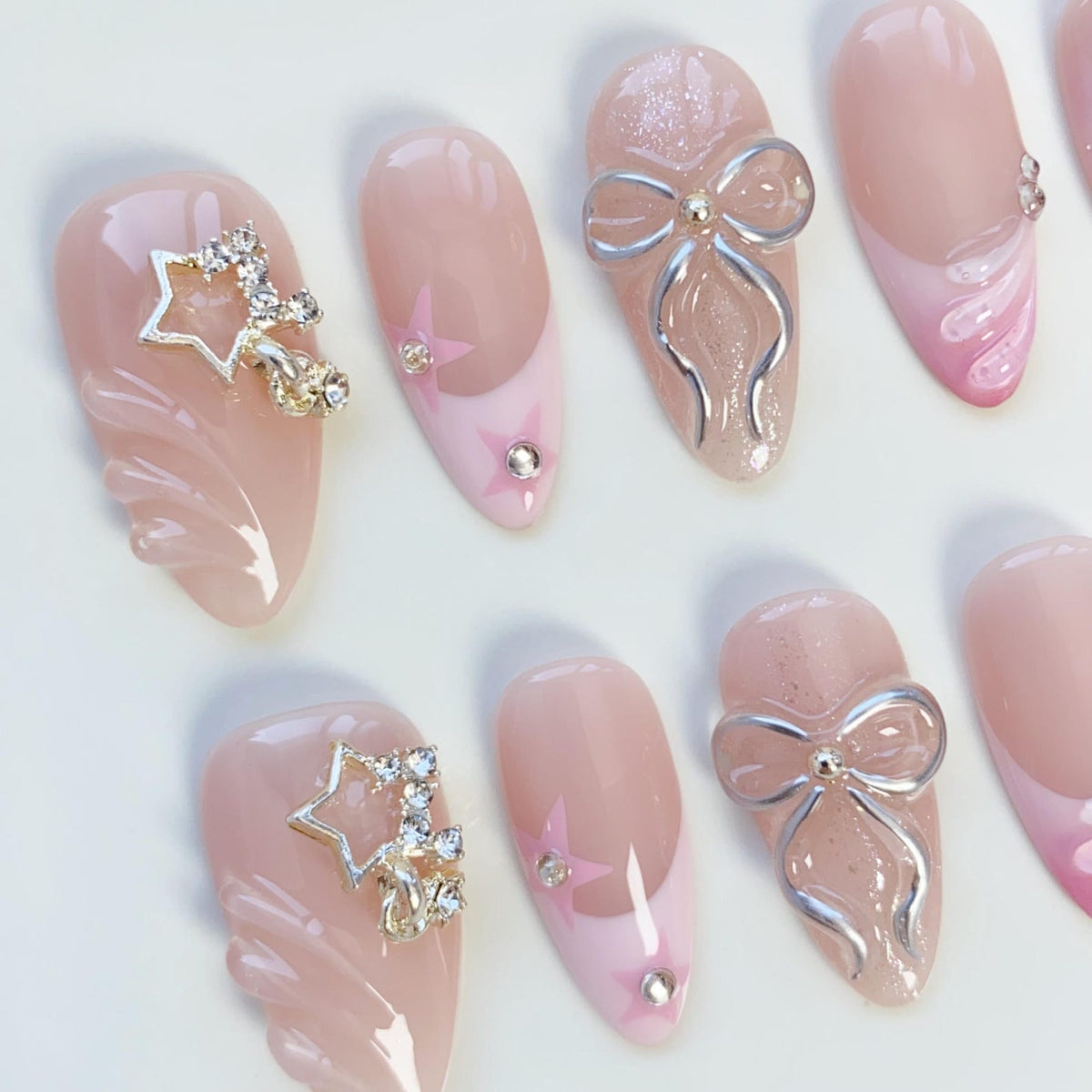 A collection of artificial nails in various designs. Some nails feature a light pink base with a star design and rhinestones. Others have a silver bow accent. The nails are oval-shaped and have a glossy finish.