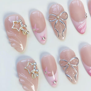 A collection of artificial nails in various designs. Some nails feature a light pink base with a star design and rhinestones. Others have a silver bow accent. The nails are oval-shaped and have a glossy finish.
