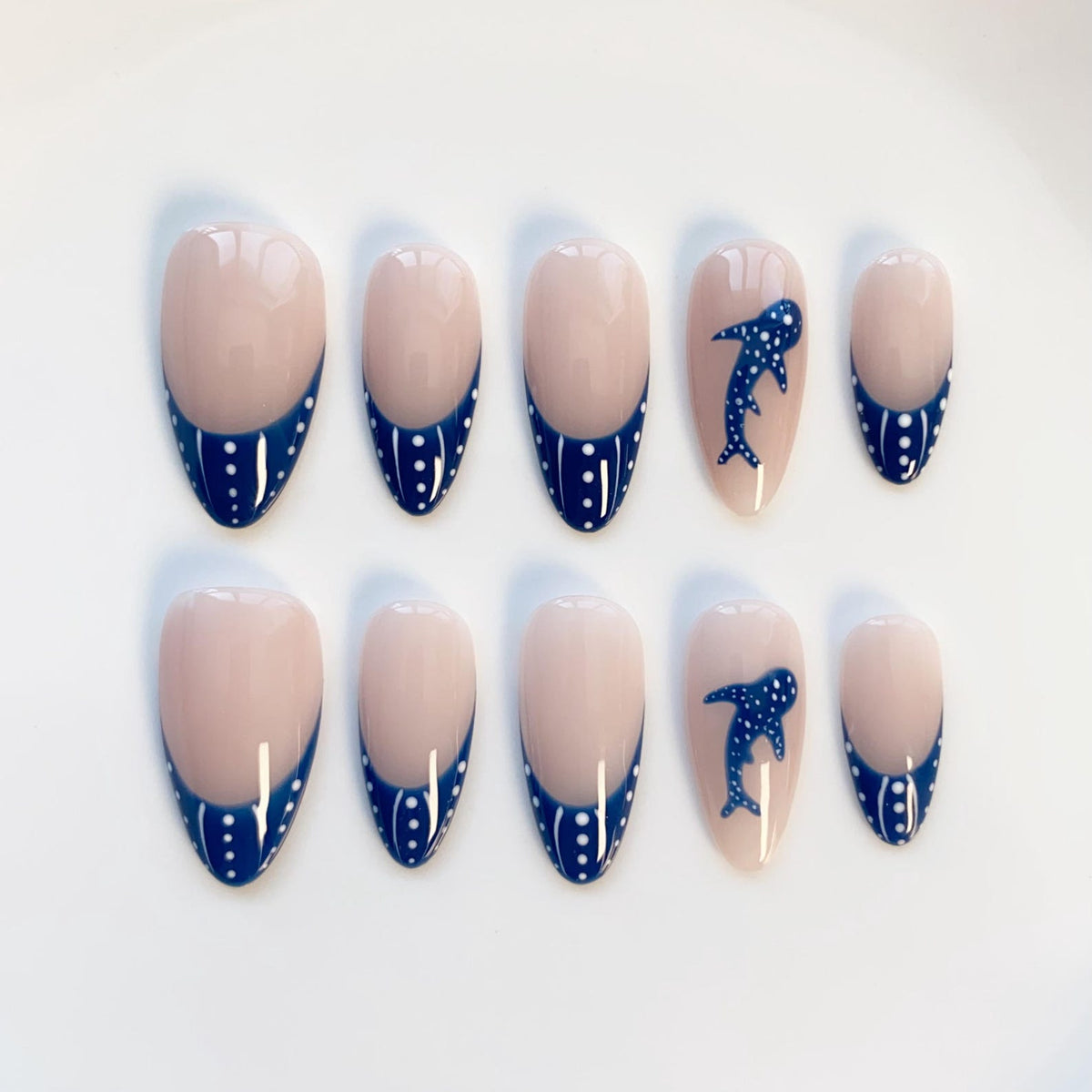 A set of ten oval-shaped false nails. The nails have a nude base with a navy blue French tip adorned with white polka dots. Two nails feature a navy blue whale design.