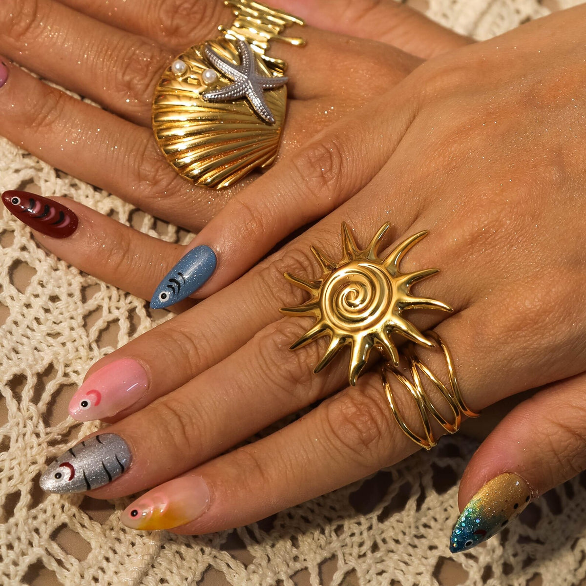 A gold-colored ring in the shape of a sun with a spiral center and spiky rays. A gold seashell ring with a starfish and pearls is also visible. The nails are painted with fish designs and ombre colors