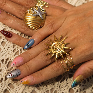 A gold-colored ring in the shape of a sun with a spiral center and spiky rays. A gold seashell ring with a starfish and pearls is also visible. The nails are painted with fish designs and ombre colors