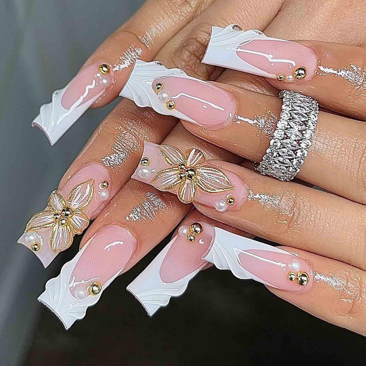 Long, sculpted artificial nails with a pink and white design. The nails feature a French tip, gold and pearl embellishments, and 3D floral accents. A silver ring with clear stones is also visible.