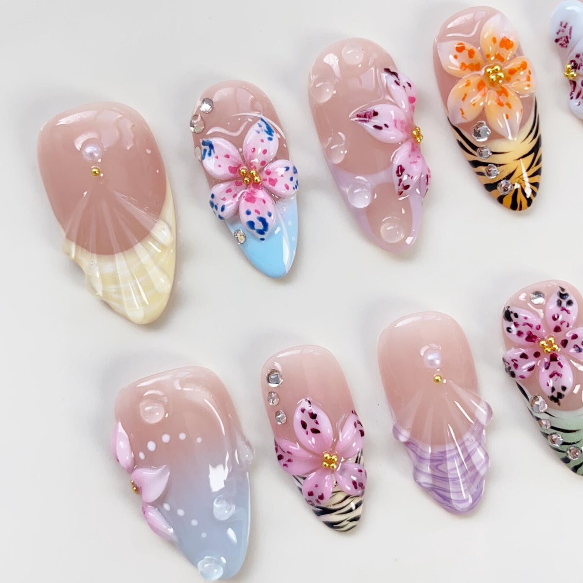 A collection of artificial nails with various designs. Some nails feature 3D floral embellishments in pink, orange, and purple, accented with rhinestones. Others have gradient color schemes and seashell-inspired patterns.