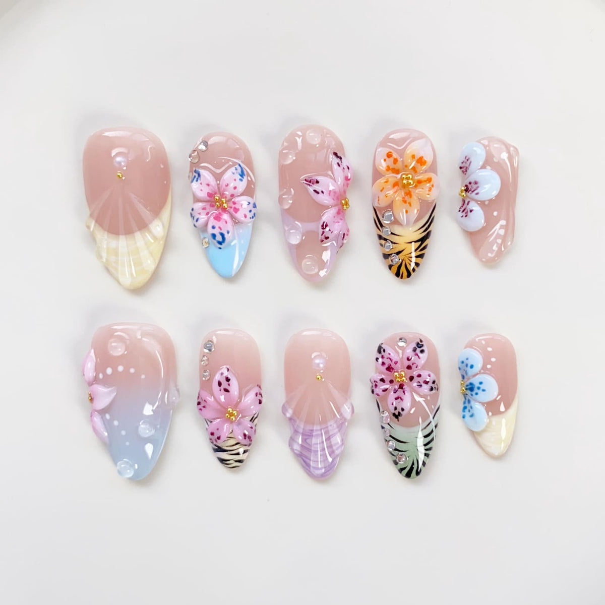 A set of ten artificial nails with a variety of floral designs. Each nail features a different design, including 3D flowers, glitter, and patterned accents. The nails are in various shades of pink, blue, and yellow.