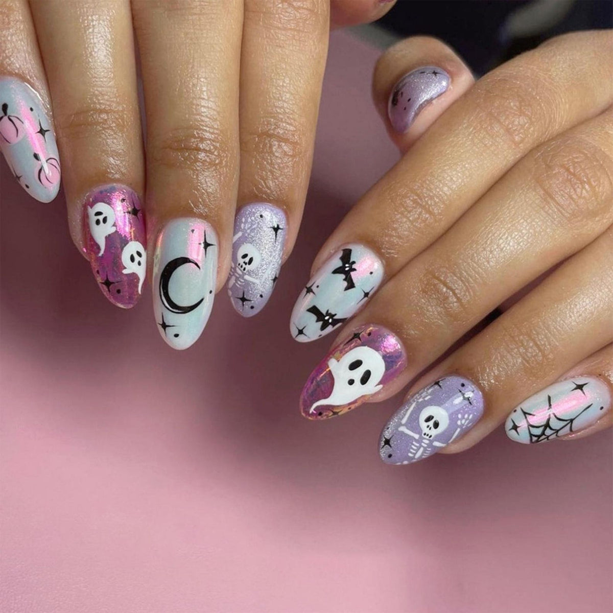 Oval-shaped nail art featuring Halloween-themed designs. The nails showcase ghosts, bats, spiders, spiderwebs, moons, and skulls in various colors, including white, purple, and pink, set against a pink background.
