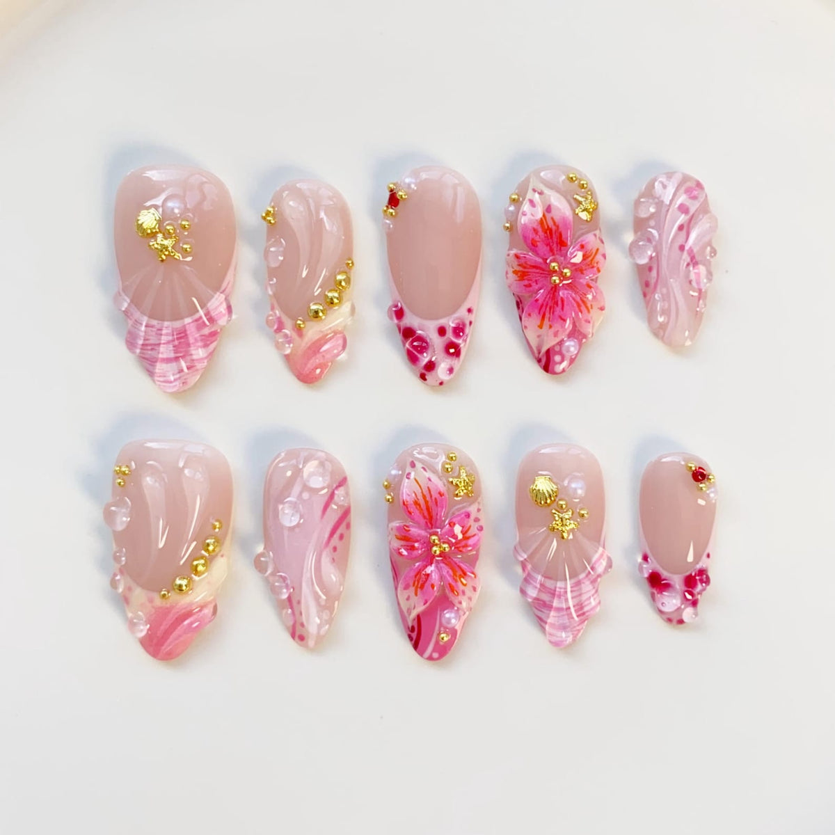 A set of ten almond-shaped artificial nails. Each nail features a nude base with pink and gold accents. Designs include floral patterns, glitter, and textured details. The nails are arranged on a white surface.