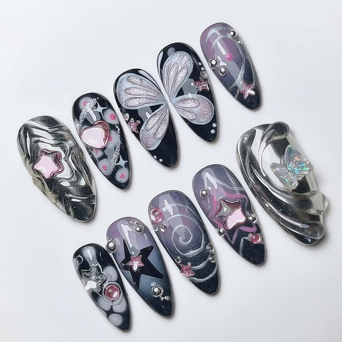 A collection of ten artificial nails with various designs. The nails feature a mix of colors including black, silver, pink, and purple. Some nails have star, heart, and butterfly designs, while others have swirls and embellishments.