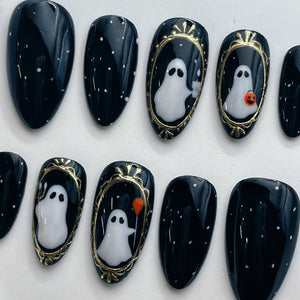 A set of press-on nails featuring a glossy black base with white speckles. Several nails display white ghost figures framed in ornate gold borders, with some ghosts holding small orange pumpkins or a 
