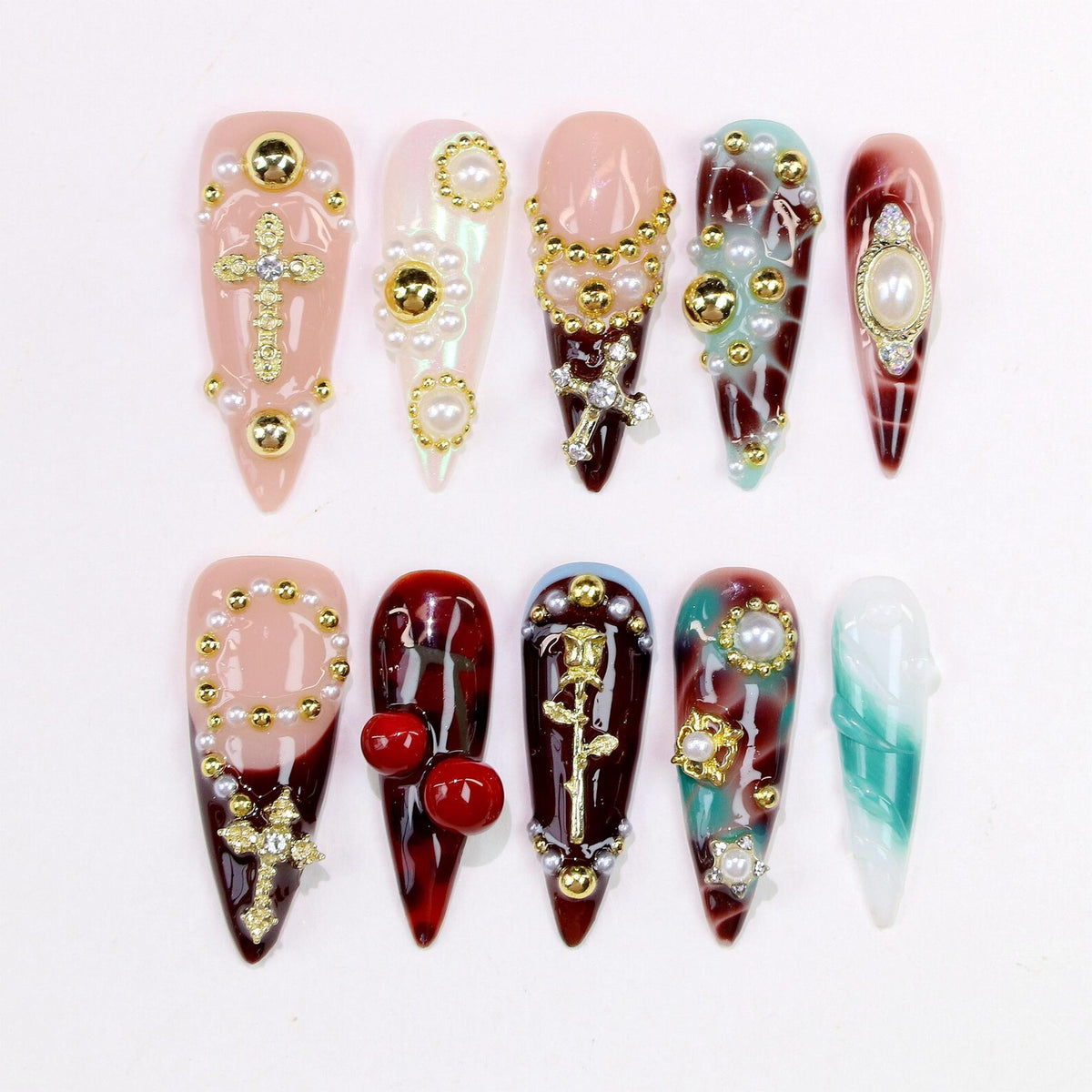 A collection of ten long, almond-shaped artificial nails. Each nail features a unique design with embellishments like pearls, crosses, and gold accents. Colors include pink, white, red, and teal, creating a glamorous aesthetic.