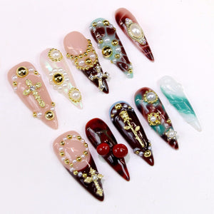 A collection of long, pointed artificial nails with various designs. Some nails feature gold crosses, pearls, and gold accents. Others have a marbled effect in shades of red, green, and white. The nails are arranged on a white surface.