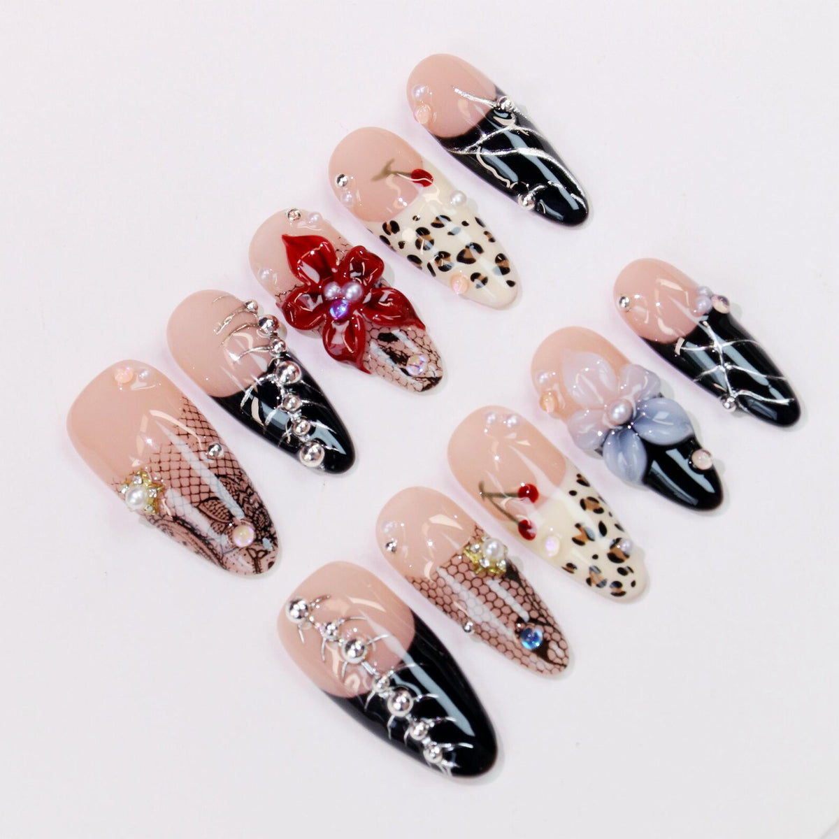 A collection of artificial nails with various designs. Some nails feature a nude base with black tips and silver accents. Others showcase leopard print, floral embellishments, and lace patterns. The nails are adorned with rhinestones and pearls.