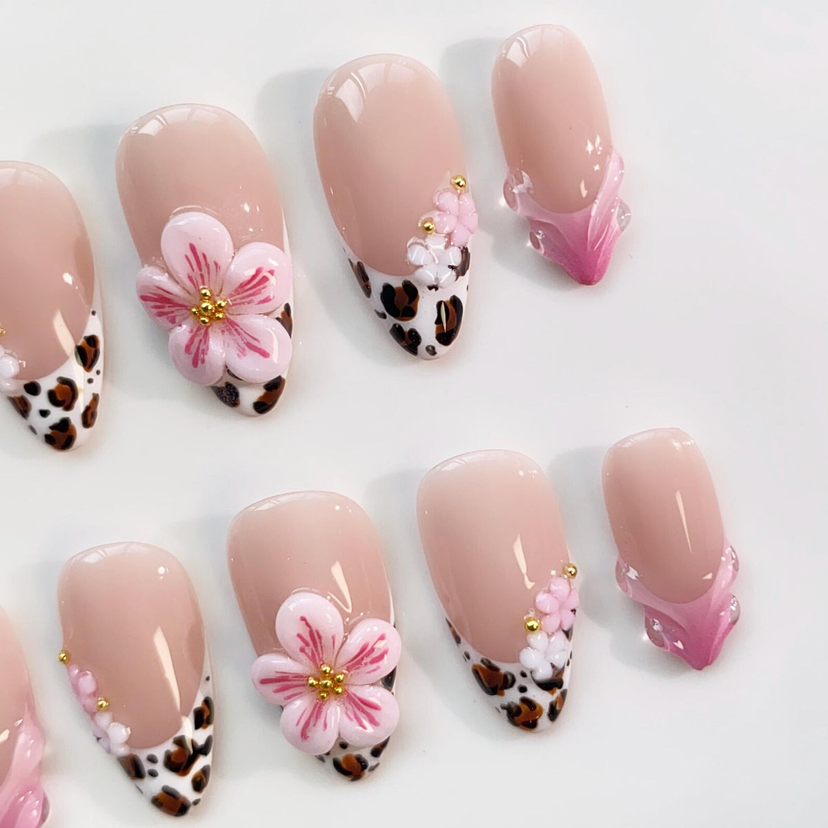 Set of artificial nails with a nude base, featuring various designs. Some nails have a leopard print tip, others have 3D pink flower accents with gold centers, and some have a pink and clear design.