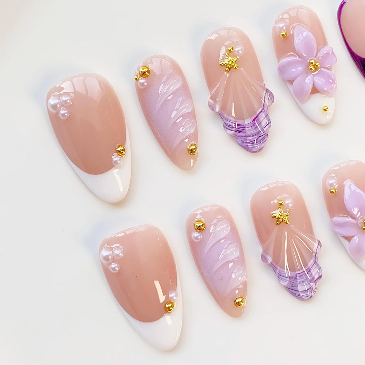 A collection of artificial nails in various designs. Some nails feature a French tip with pearl and gold accents. Others showcase a swirl design in shades of pink and purple, and some have 3D flower and seashell embellishments.