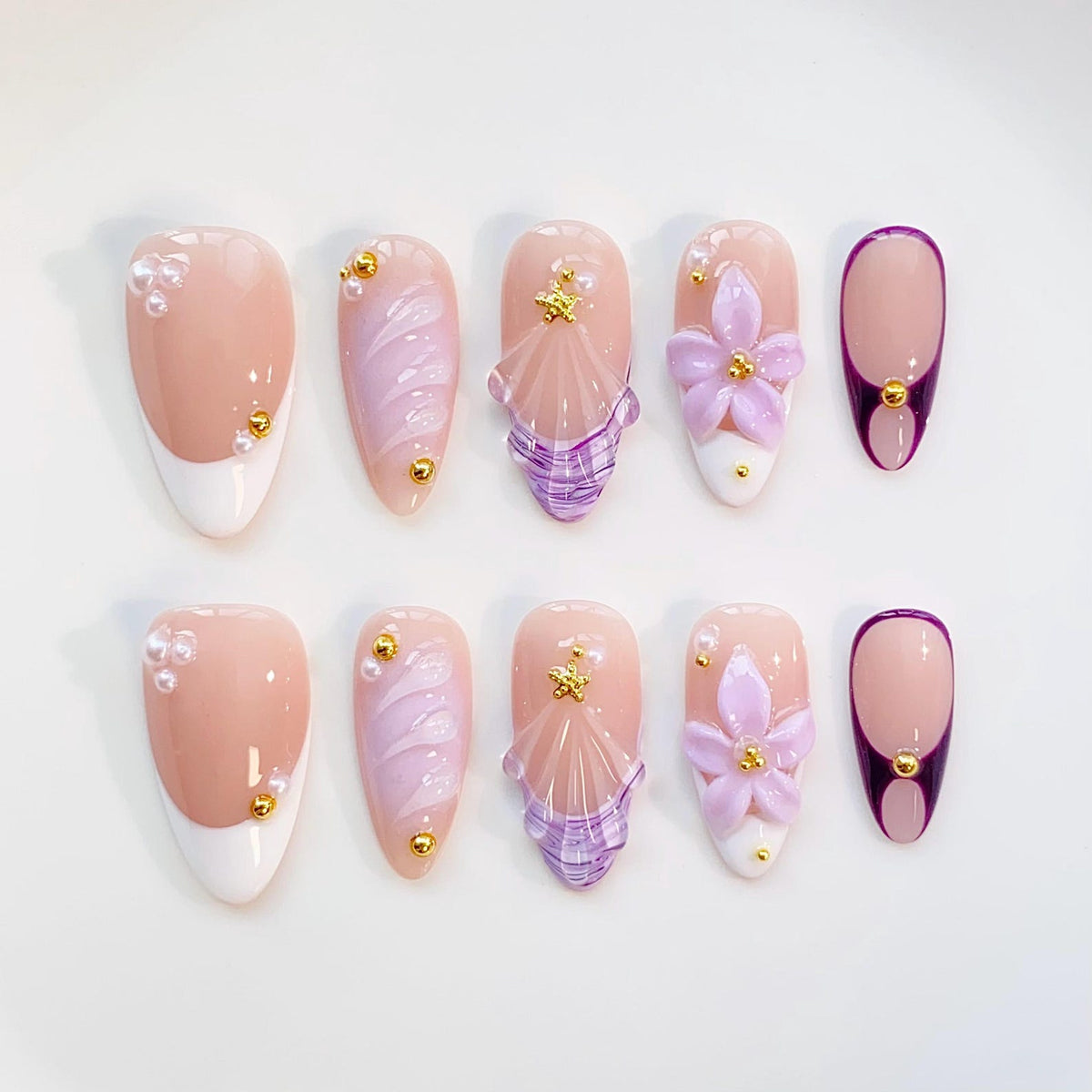 A set of ten artificial nails in various designs. The nails feature a light pink base with white tips, lavender swirls, and 3D floral and seashell accents. Some nails have pearl and gold embellishments.