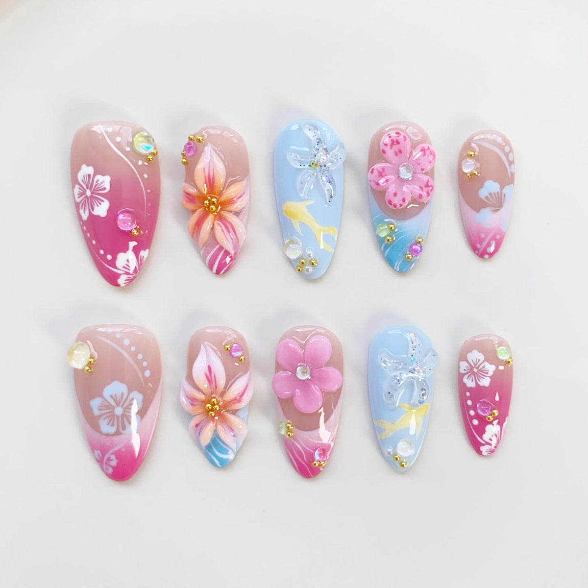 A set of ten artificial nails with a variety of floral and aquatic designs. Each nail features a different design, including pink and white flowers, 3D flowers, and aquatic elements like starfish and fish. The nails are painted in shades of pink, blu