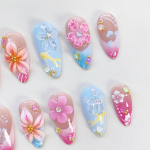 A collection of almond-shaped artificial nails with various designs. Some nails feature 3D pink and orange flowers with gold accents. Others have blue backgrounds with starfish, fish, and clear gems. The nails have a glossy finish.