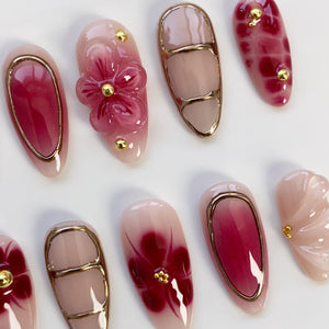A collection of artificial nails with various designs. Some nails feature a gradient of pink and red hues, while others showcase floral embellishments and gold accents. The nails are oval-shaped and have a glossy finish.