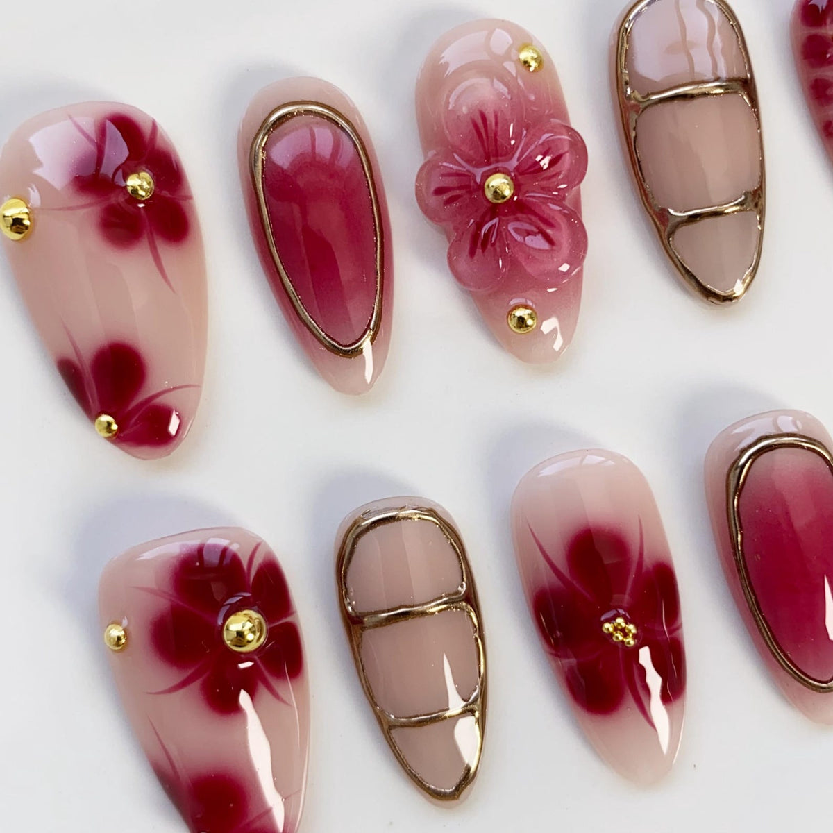 A collection of artificial nails with various designs. Some nails feature a gradient of pink and burgundy, adorned with gold accents and floral patterns. Others showcase a 3D flower design and gold borders, creating a sophisticated and elegant aesthe