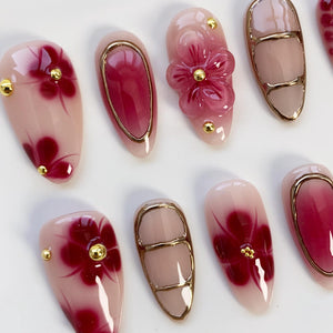 A collection of artificial nails with various designs. Some nails feature a gradient of pink and burgundy, adorned with gold accents and floral patterns. Others showcase a 3D flower design and gold borders, creating a sophisticated and elegant aesthe