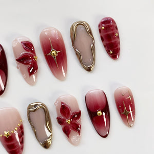 A collection of artificial nails in various designs. Some nails feature a gradient of pink to burgundy, others have gold starbursts, floral accents, or gold embellishments. The nails are oval-shaped and have a glossy finish.