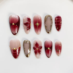 A set of ten artificial nails in various designs. The nails feature a gradient of red and pink hues, with gold accents, floral embellishments, and geometric shapes. Some nails have a glossy finish, while others have a matte appearance.