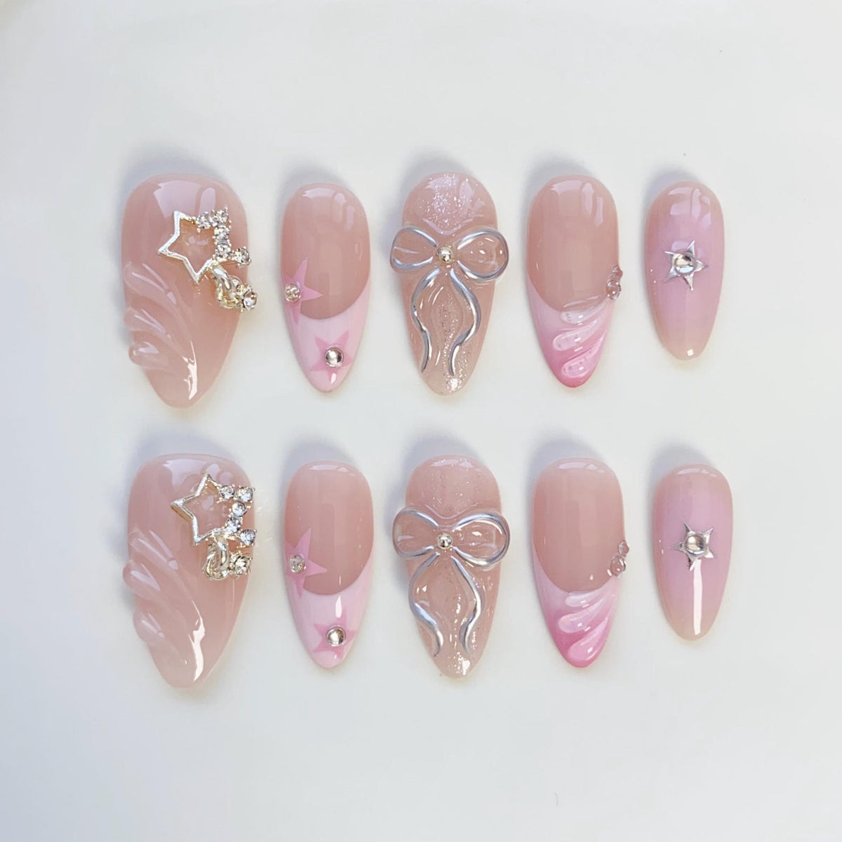 A set of ten artificial nails in various designs. The nails are primarily light pink with accents of silver bows, star-shaped rhinestones, and pink swirls. Some nails feature a French tip design.