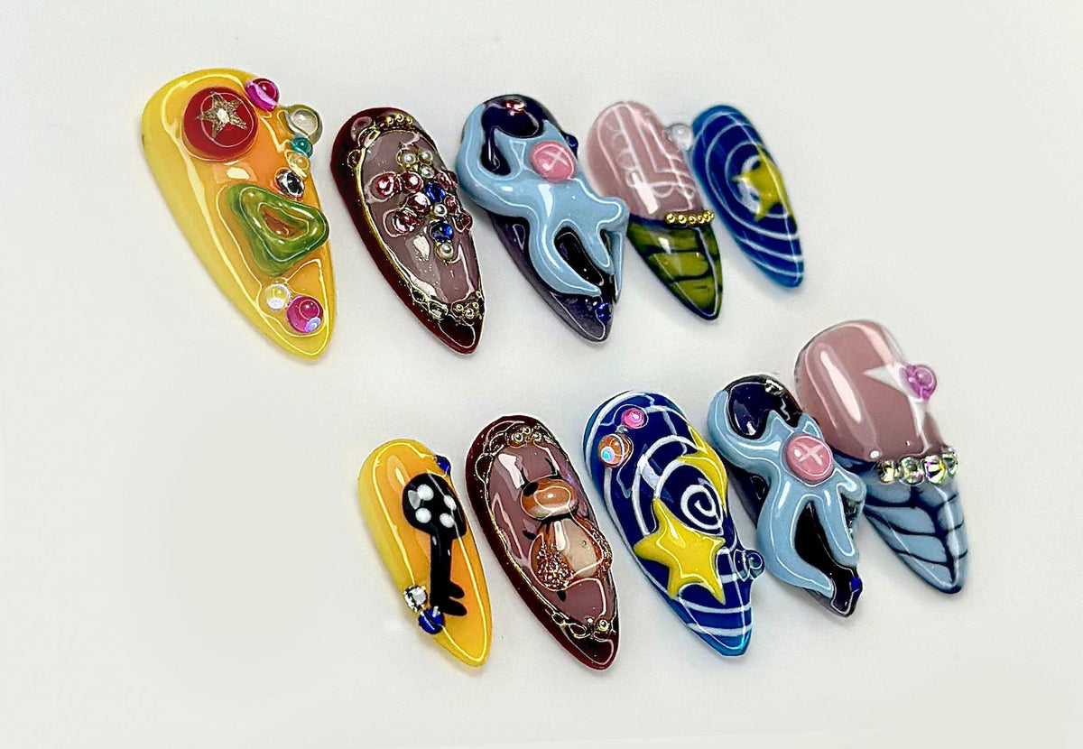 A collection of ten almond-shaped, embellished artificial nails. Designs include a key, an octopus, and a star, with colors like yellow, blue, and brown. Each nail is adorned with gems and intricate details.
