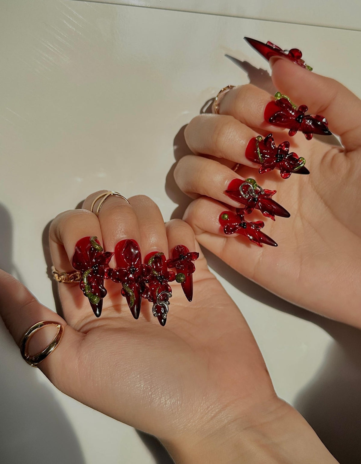 A close-up of two hands with long, red acrylic nails. Each nail features a dark red, pointed tip and is adorned with intricate, dark red floral designs with green and silver accents. Gold rings are visible on the fingers.