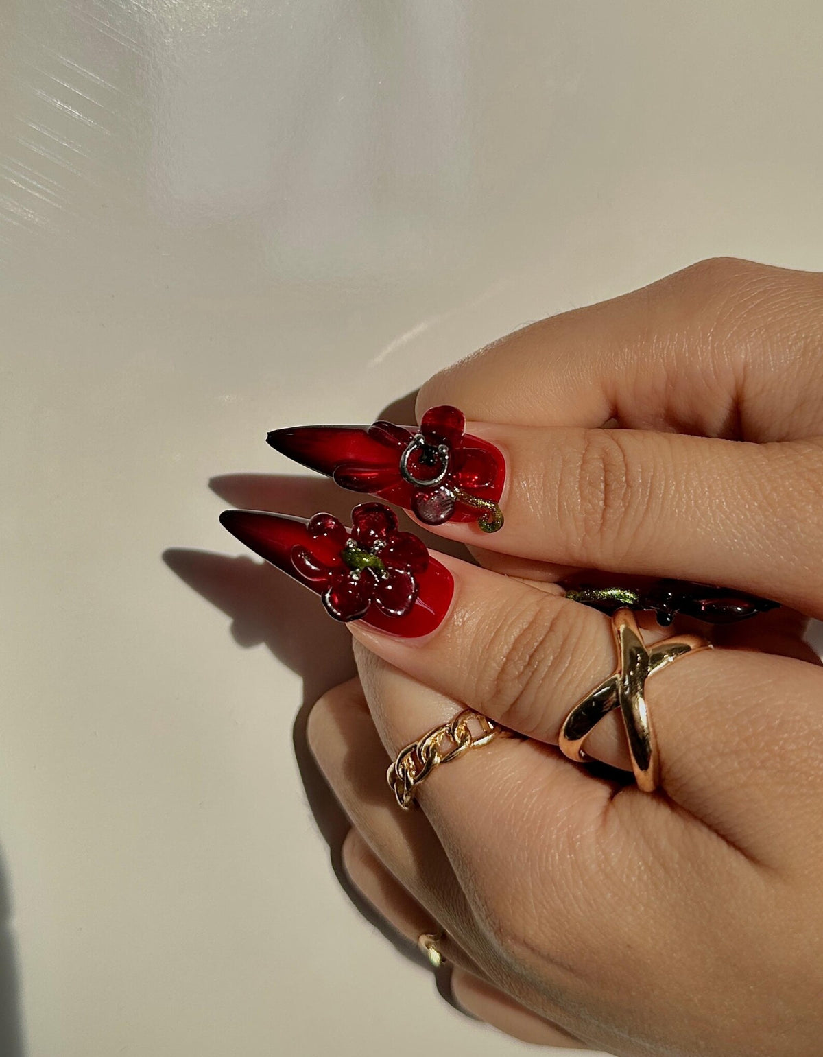 Close-up of red stiletto nails with 3D flower and gem designs. The nails are adorned with gold rings, including a chain link and a crisscross design. The nails have a glossy finish.