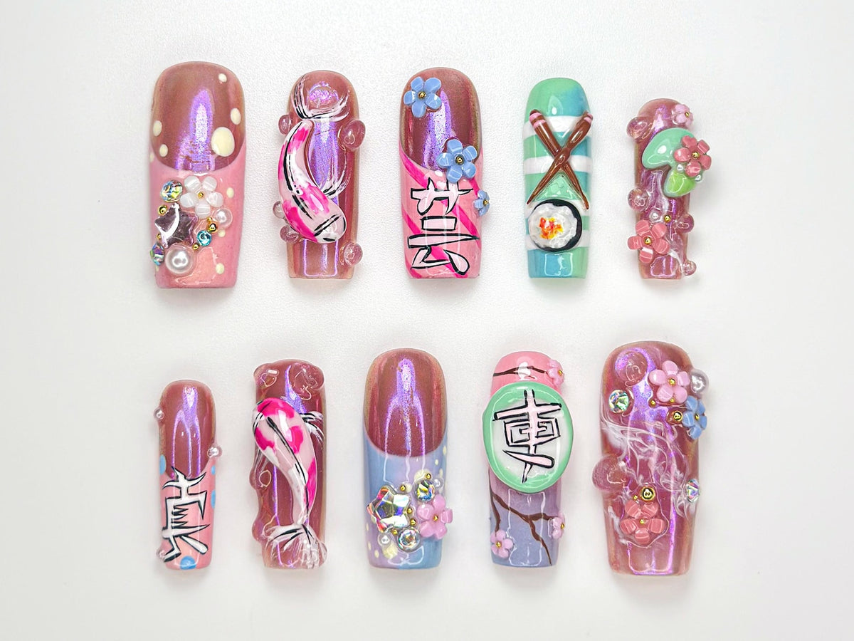 A set of ten artificial nails with various designs. The nails feature a range of colors, including pink, purple, and green, and are adorned with embellishments such as flowers, koi fish, and Japanese characters. Some nails have a glossy finish.