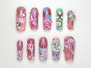 A set of ten artificial nails with various designs. The nails feature a range of colors, including pink, purple, and green, and are adorned with embellishments such as flowers, koi fish, and Japanese characters. Some nails have a glossy finish.