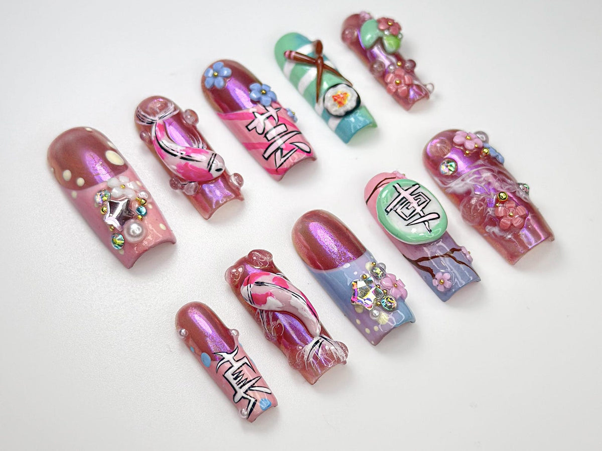 A collection of ten artificial nails with intricate designs. The nails feature a range of colors, including pink, purple, blue, and green, with detailed artwork such as flowers, koi fish, and Japanese characters. Some nails have added embellishments.